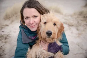Eine Person mit langem Haar umarmt während einer Kinderhypnosesitzung einen flauschigen Hund an einem Sandstrand. Die Person trägt eine blaugrüne Jacke und einen Schal, während der Hund hellbraun ist und ein zotteliges Fell hat. Im Hintergrund sind Sand und verstreutes trockenes Gras zu sehen, was eine Atmosphäre der Ruhe schafft.