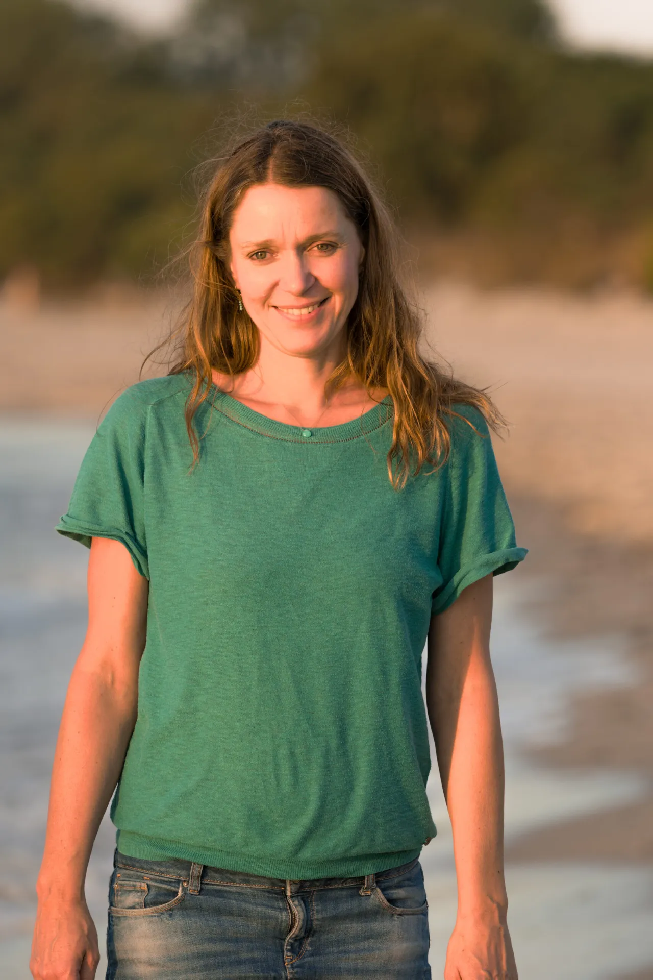 Eine Frau mit langen Haaren steht lächelnd an einem Strand und trägt ein grünes Hemd und Jeans. Im Hintergrund sind Bäume und Sand zu sehen. Die Beleuchtung lässt vermuten, dass es um Sonnenuntergang ist.