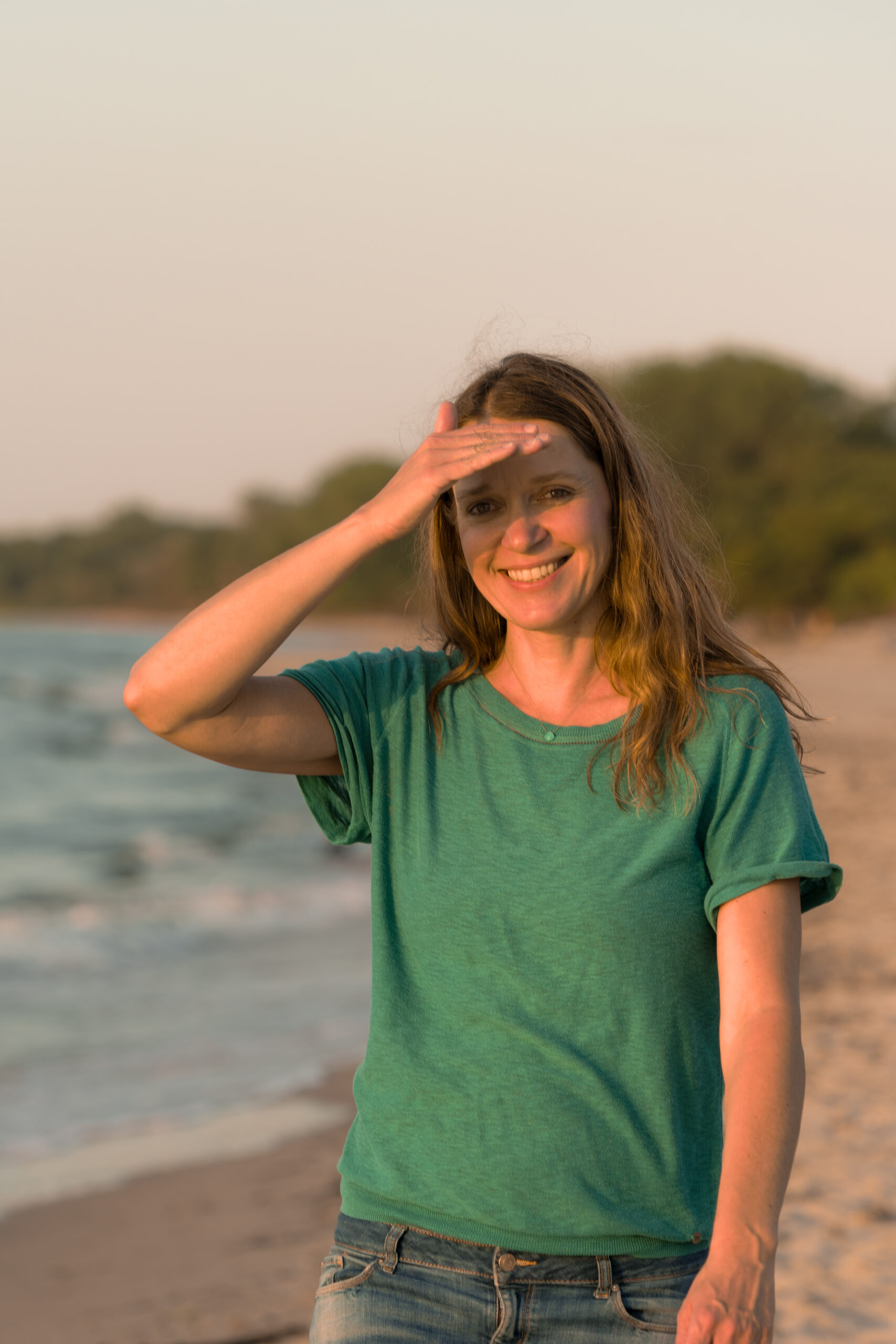 Eine Person mit langen Haaren steht lächelnd an einem Sandstrand. Sie trägt ein grünes T-Shirt und Jeans. Eine Hand ist an die Stirn gelegt, um die Augen vor der Sonne zu schützen. Im Hintergrund sind das Meer und Bäume zu sehen.