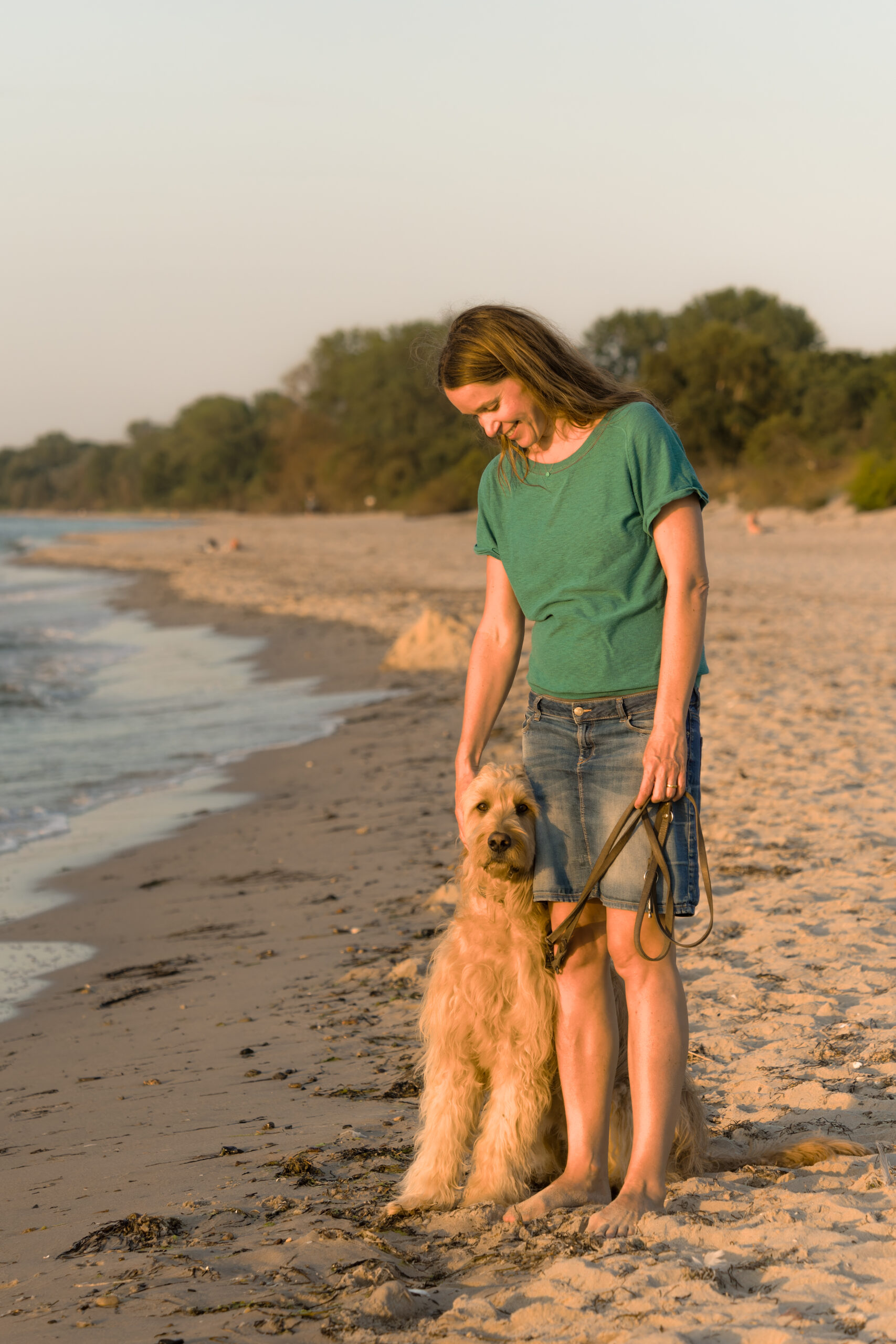 Eine Frau mit langen Haaren, einem grünen T-Shirt und Jeansshorts, steht barfuß an einem Sandstrand. Sie lächelt auf einen flauschigen, braunen Hund herab, der an der Leine neben ihr sitzt. Im Hintergrund sind ruhiges Wasser und in der Ferne Bäume unter einem klaren Himmel zu sehen.
